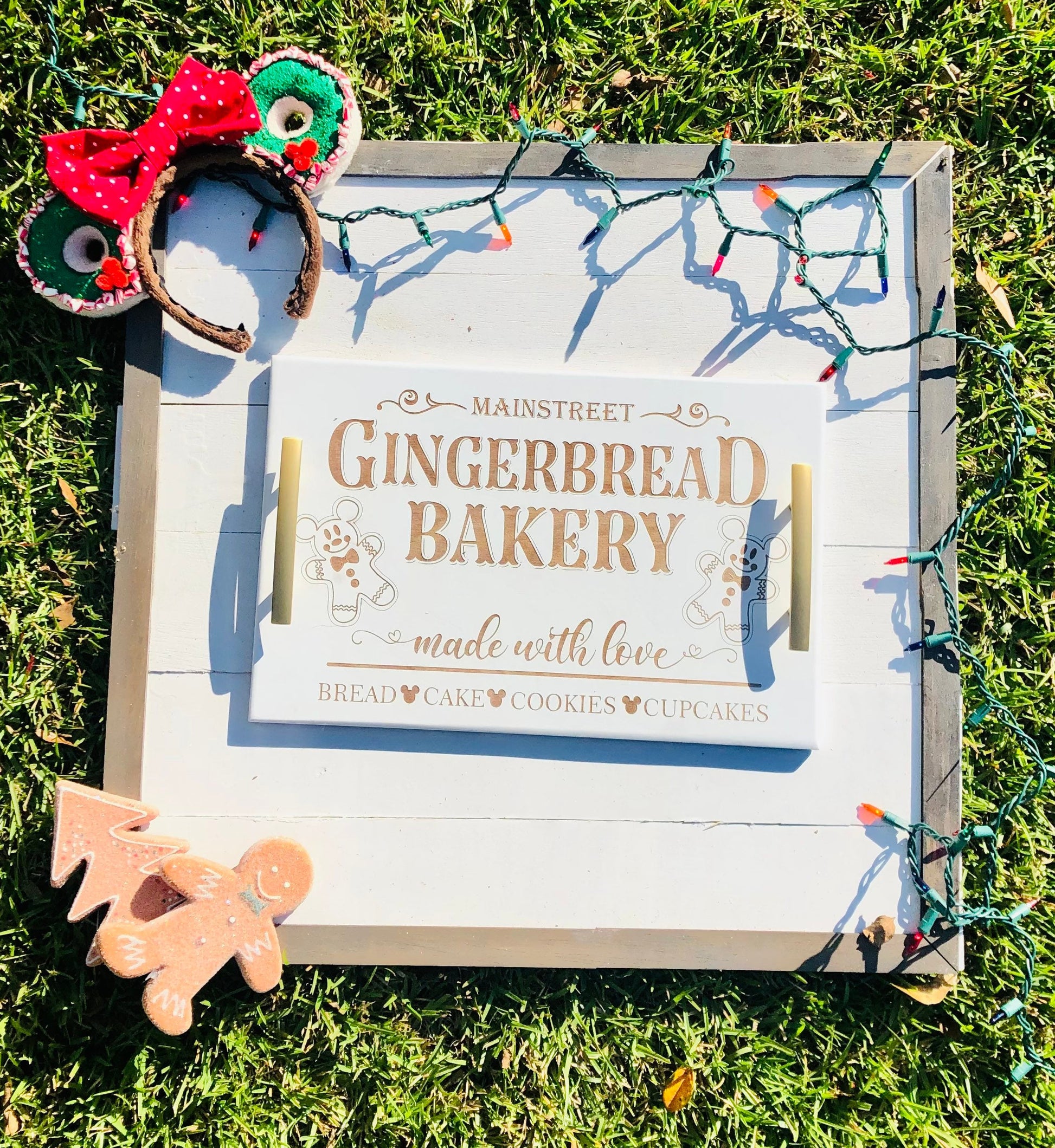 Wooden sign with 'Mainstreet Gingerbread Bakery' text, surrounded by Christmas lights and gingerbread decorations on grass.
