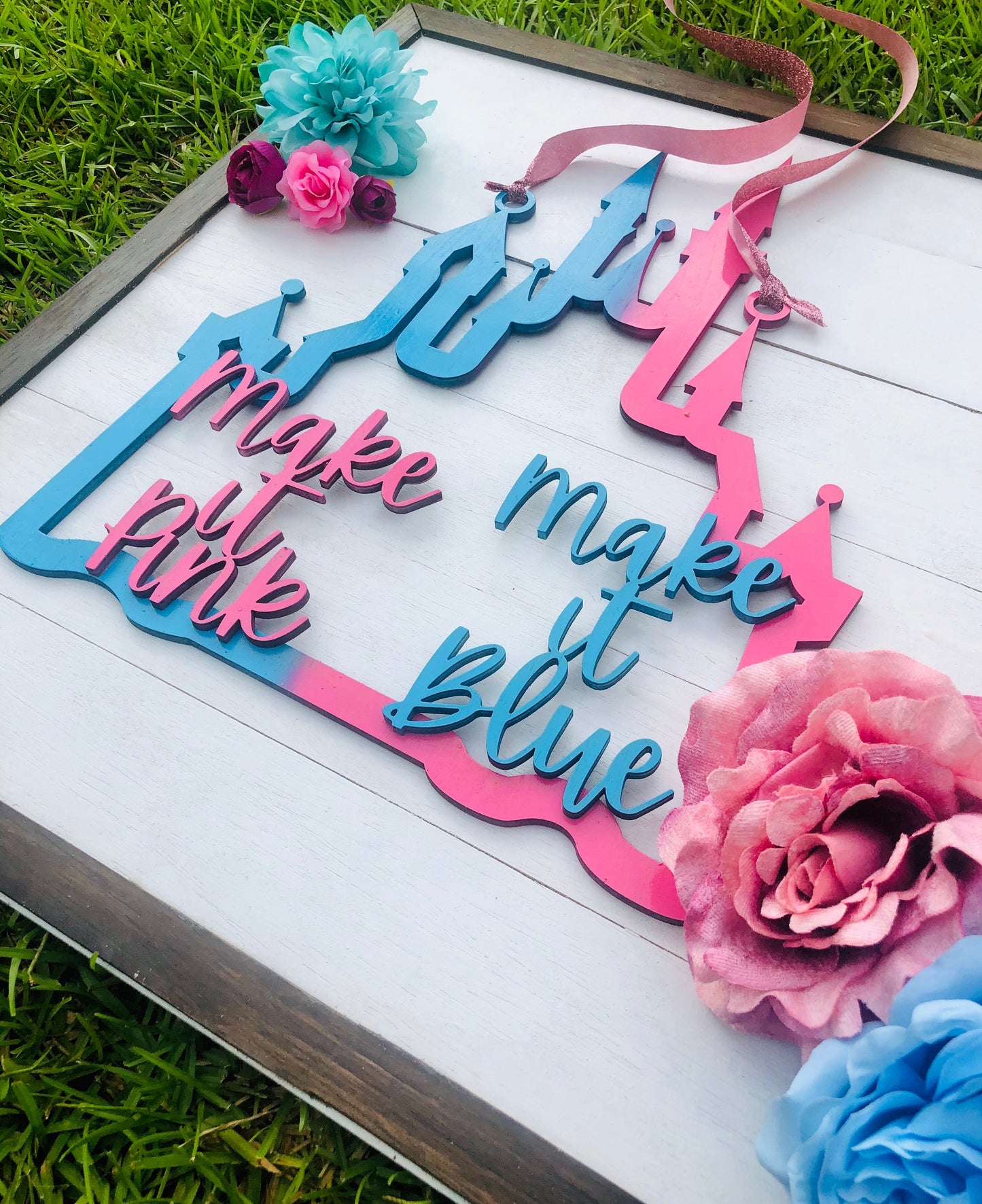 Decorative sign with 'make it pink' and 'make it blue' text, surrounded by flowers on a grass background.