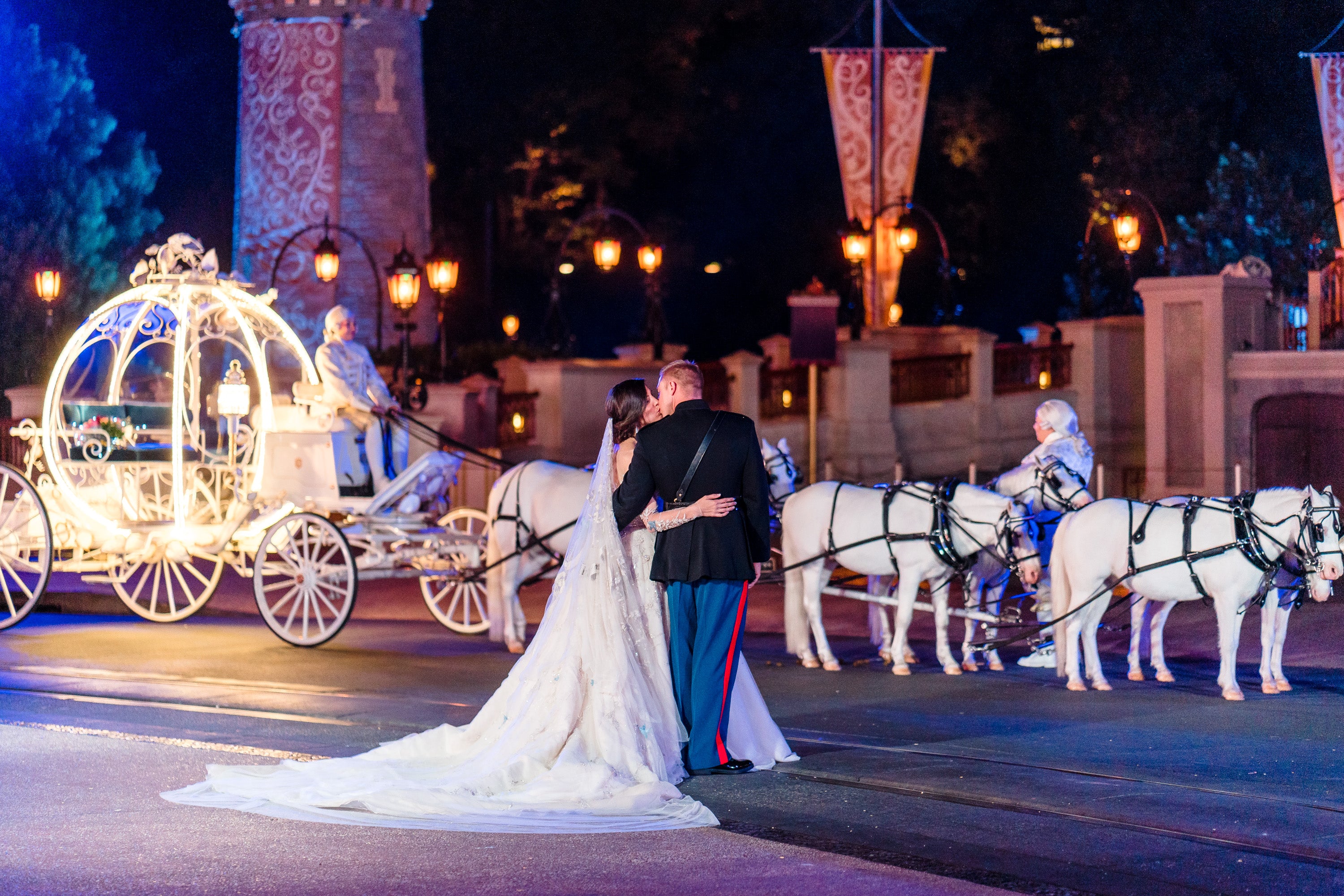 Romantic wedding couple standing arm in arm in front of a glowing Cinderella-style carriage pulled by white horses at night.