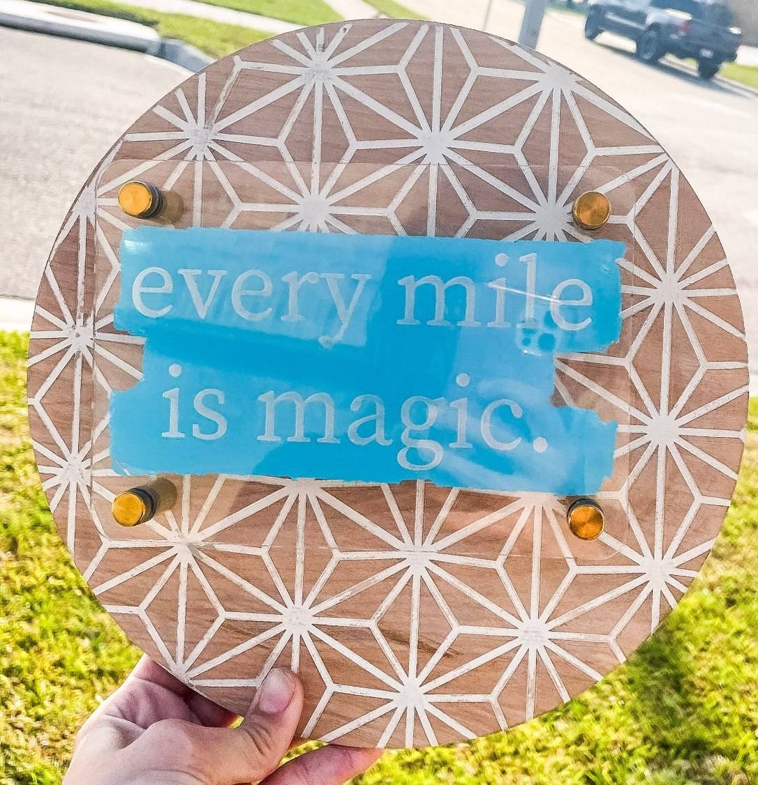Round wooden sign with geometric pattern and blue sticker saying 'every mile is magic.' held outdoors.