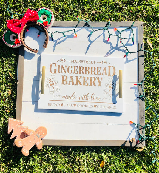 Wooden sign with 'Mainstreet Gingerbread Bakery' text, surrounded by Christmas lights and gingerbread decorations on grass.