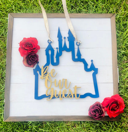Decorative sign with blue castle design and 'Be Our Guest' text, surrounded by red and purple roses on a grass background.
