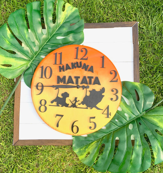 Round clock with 'Hakuna Matata' text and characters on a white background with green leaves.