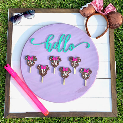 Decorative sign with 'hello' in teal, chocolate cake pops with pink bows, and a pink straw on a grass background.