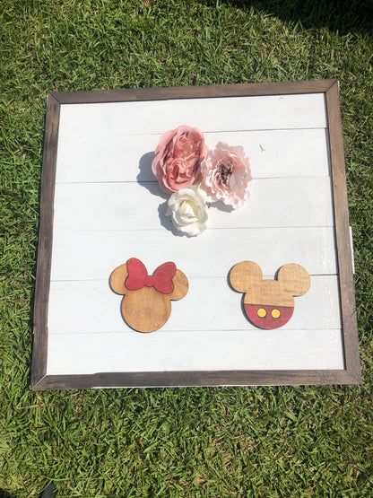Wooden earrings shaped like Mickey and Minnie Mouse heads on a white background with flowers, framed in brown.