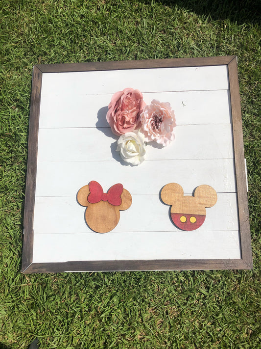Wooden earrings shaped like Mickey and Minnie Mouse heads on a white background with flowers, framed in brown.