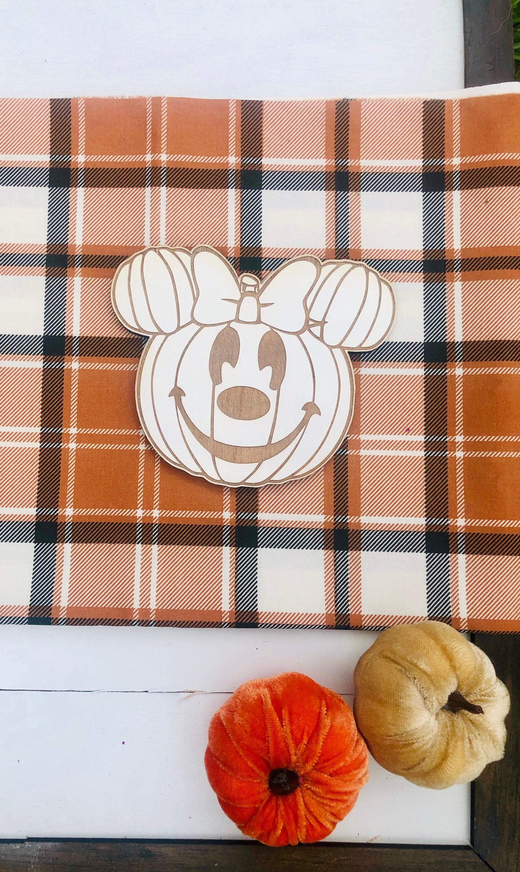 Pumpkin-shaped cutout with Minnie Mouse design on a plaid fabric background with small pumpkins.