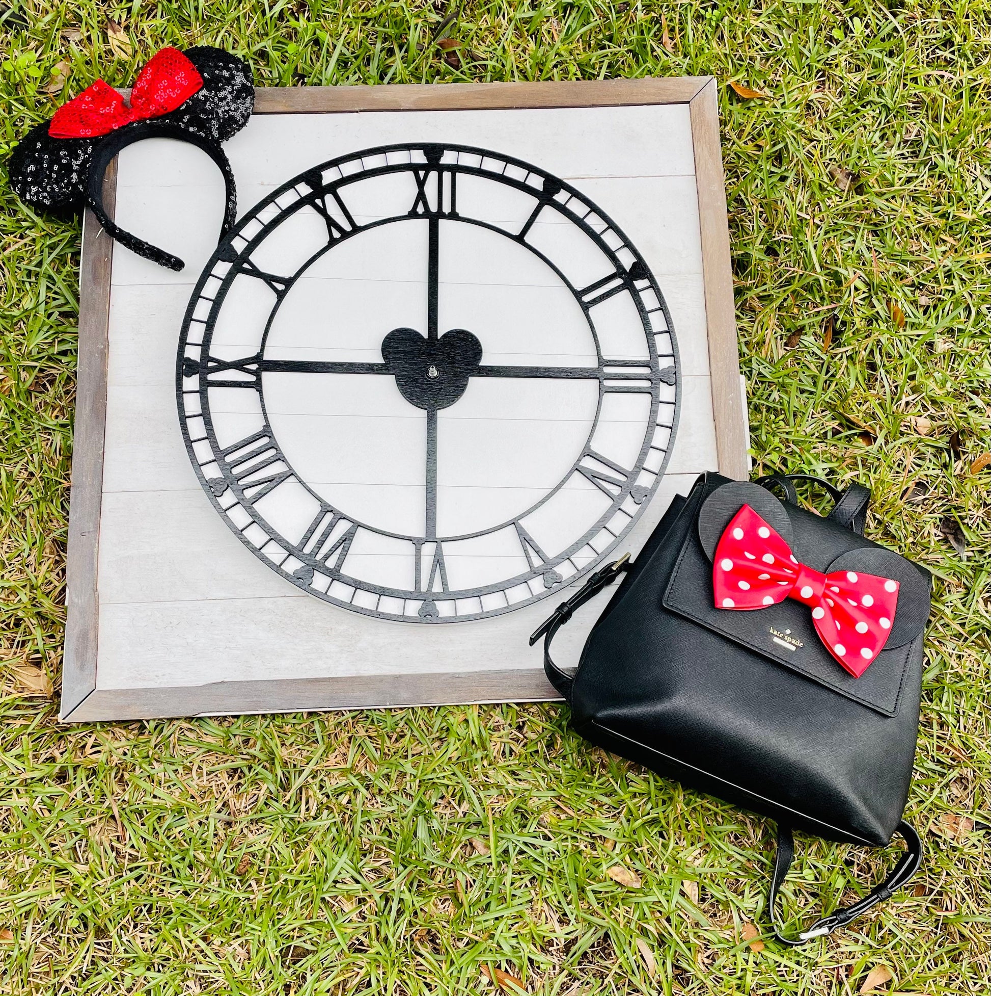Black backpack with red polka dot bow next to a clock and Minnie Mouse ears on grass