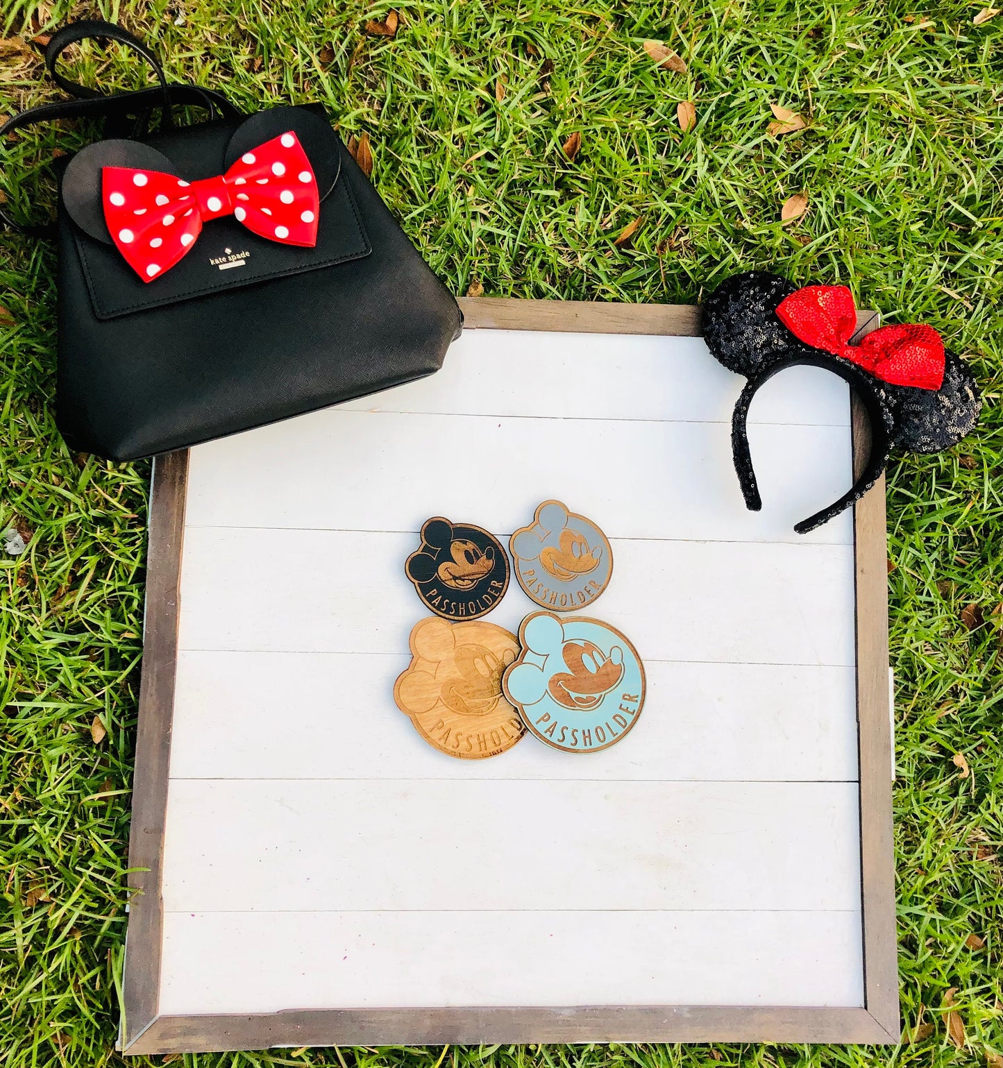 Black handbag with red polka dot bow, black mouse ears with red bow, and three Disney pins on a white surface with grass background.