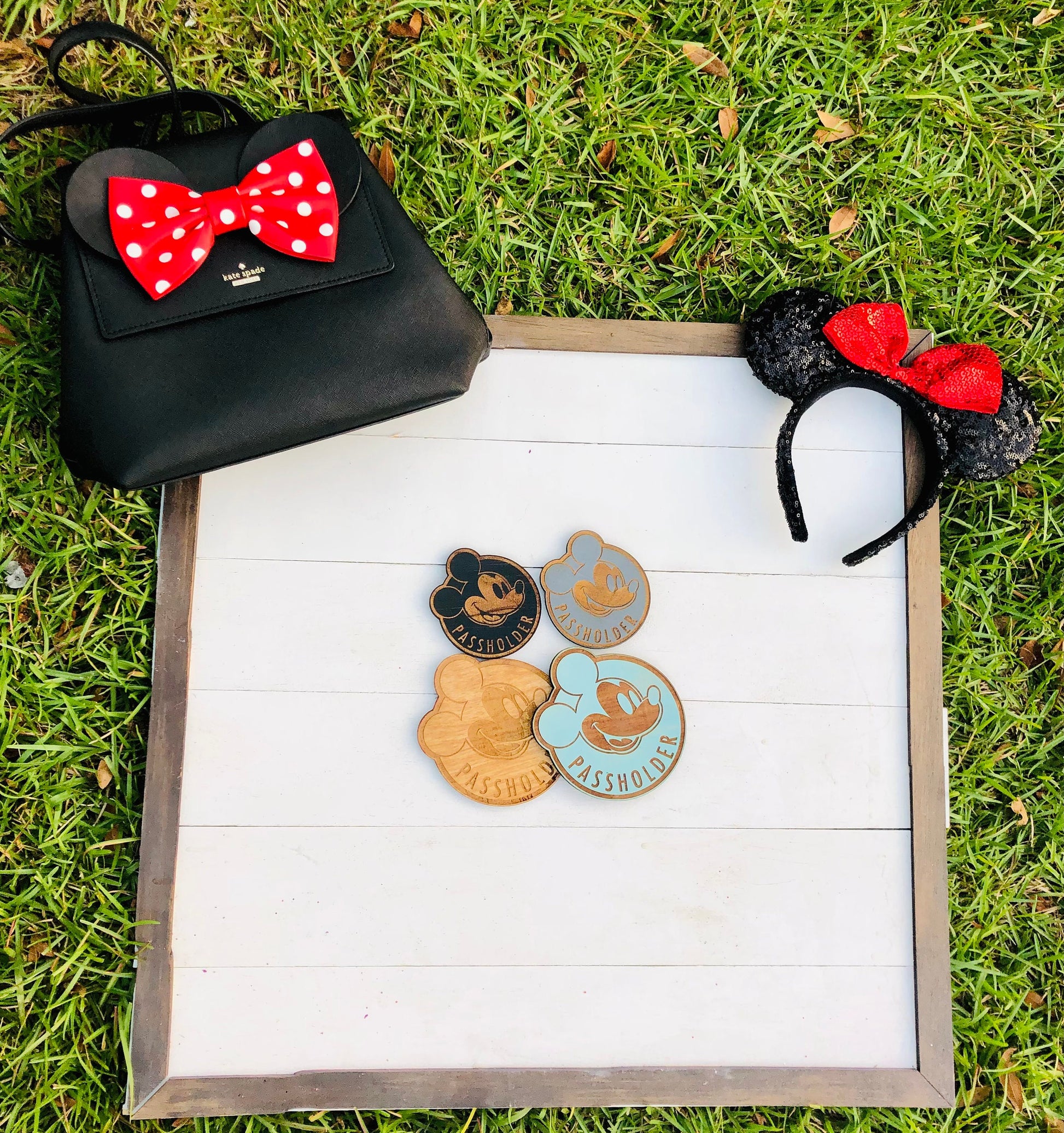 Black handbag with red polka dot bow, black mouse ears with red bow, and three Disney pins on a white surface with grass background.