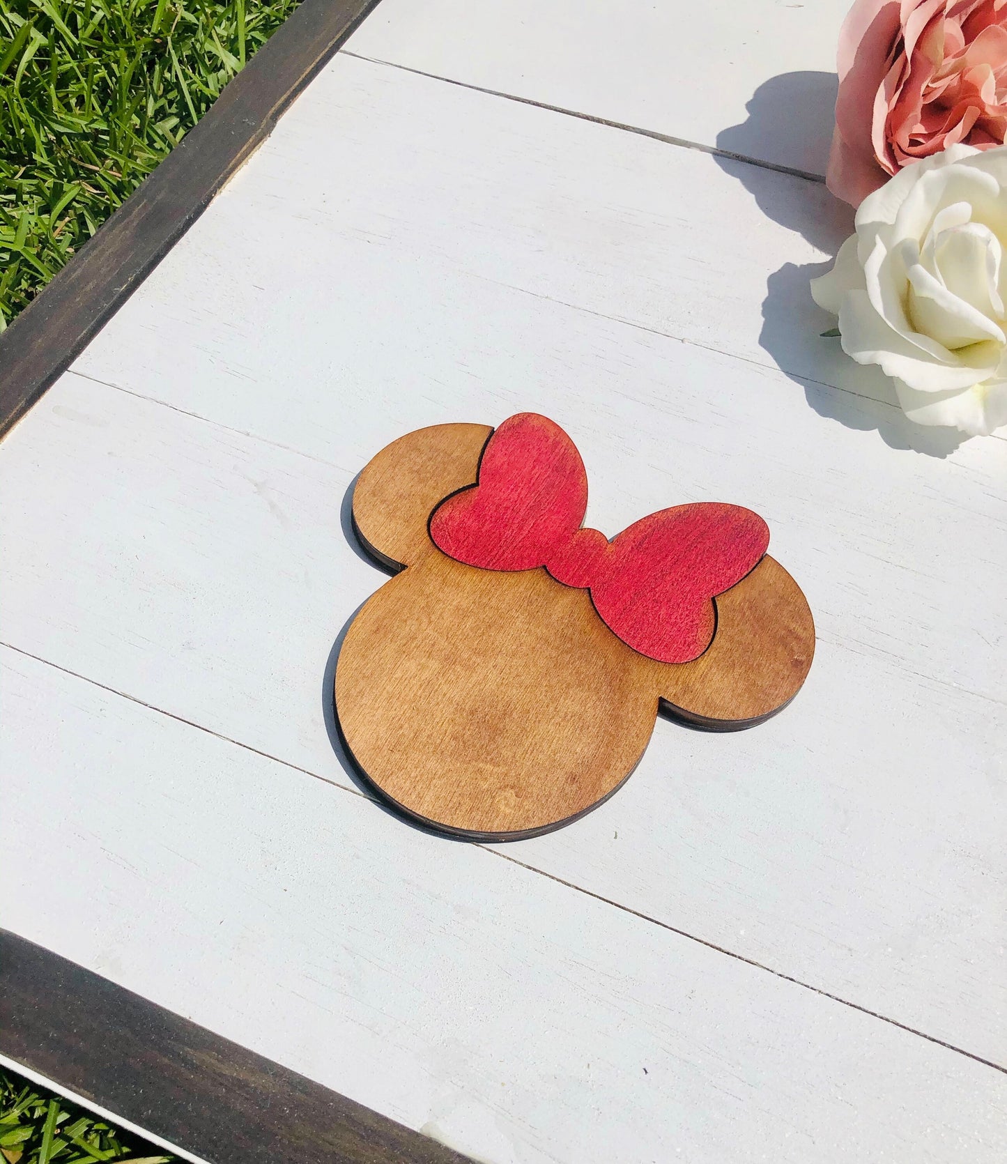 Wooden cutout of a mouse head with a red bow on a wooden surface with flowers in the background.
