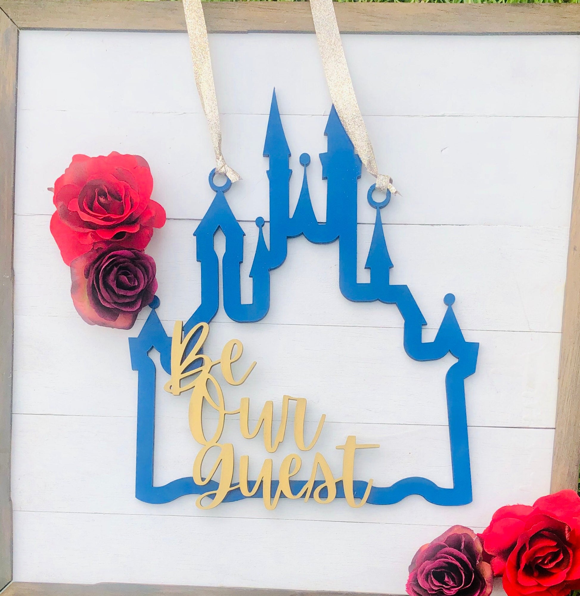 Decorative sign with blue castle design and 'Be Our Guest' text, surrounded by red and pink flowers on a white background.