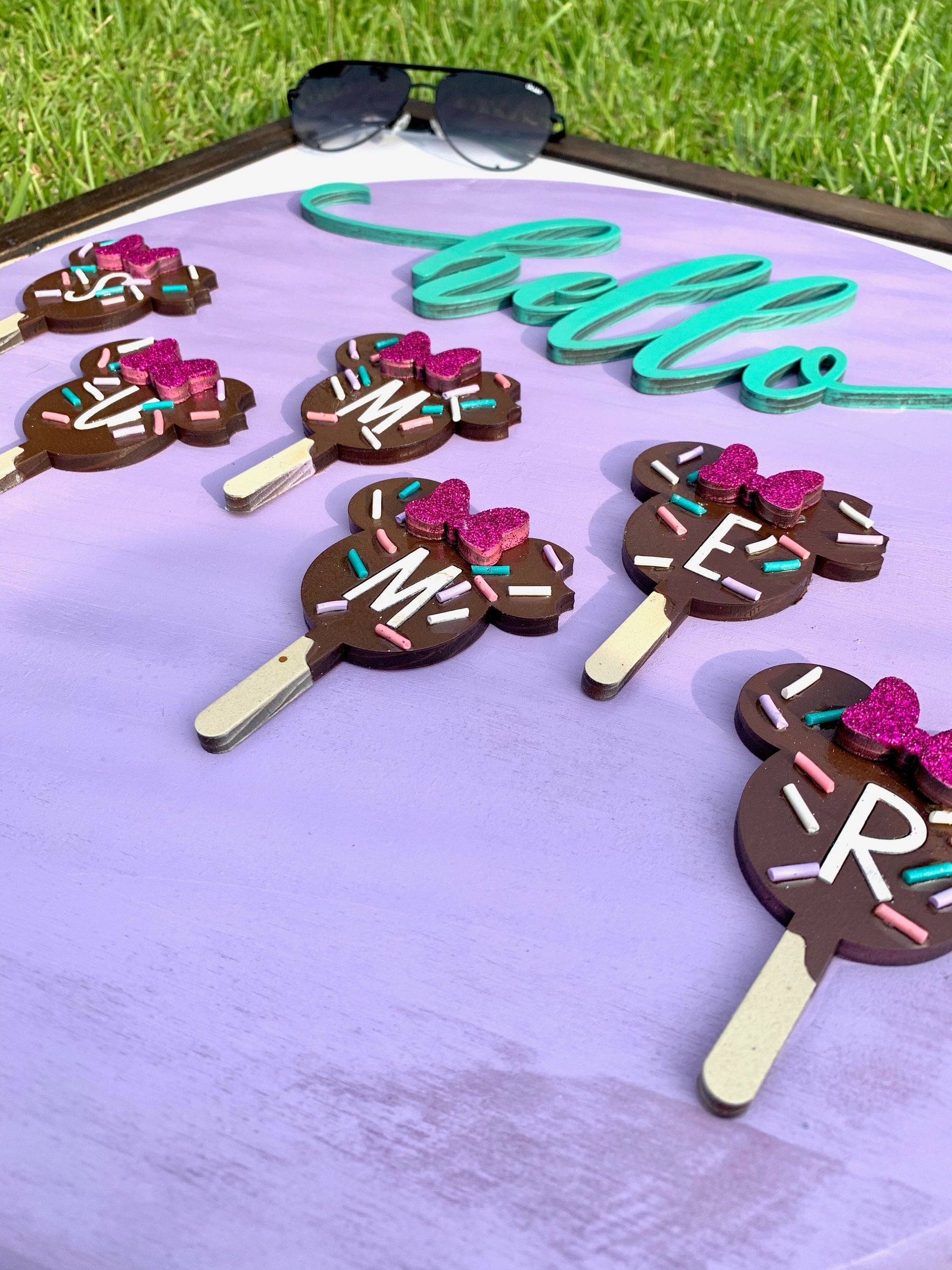Chocolate treats shaped like ice cream bars with letters on sticks, placed on a purple surface with sunglasses in the background.