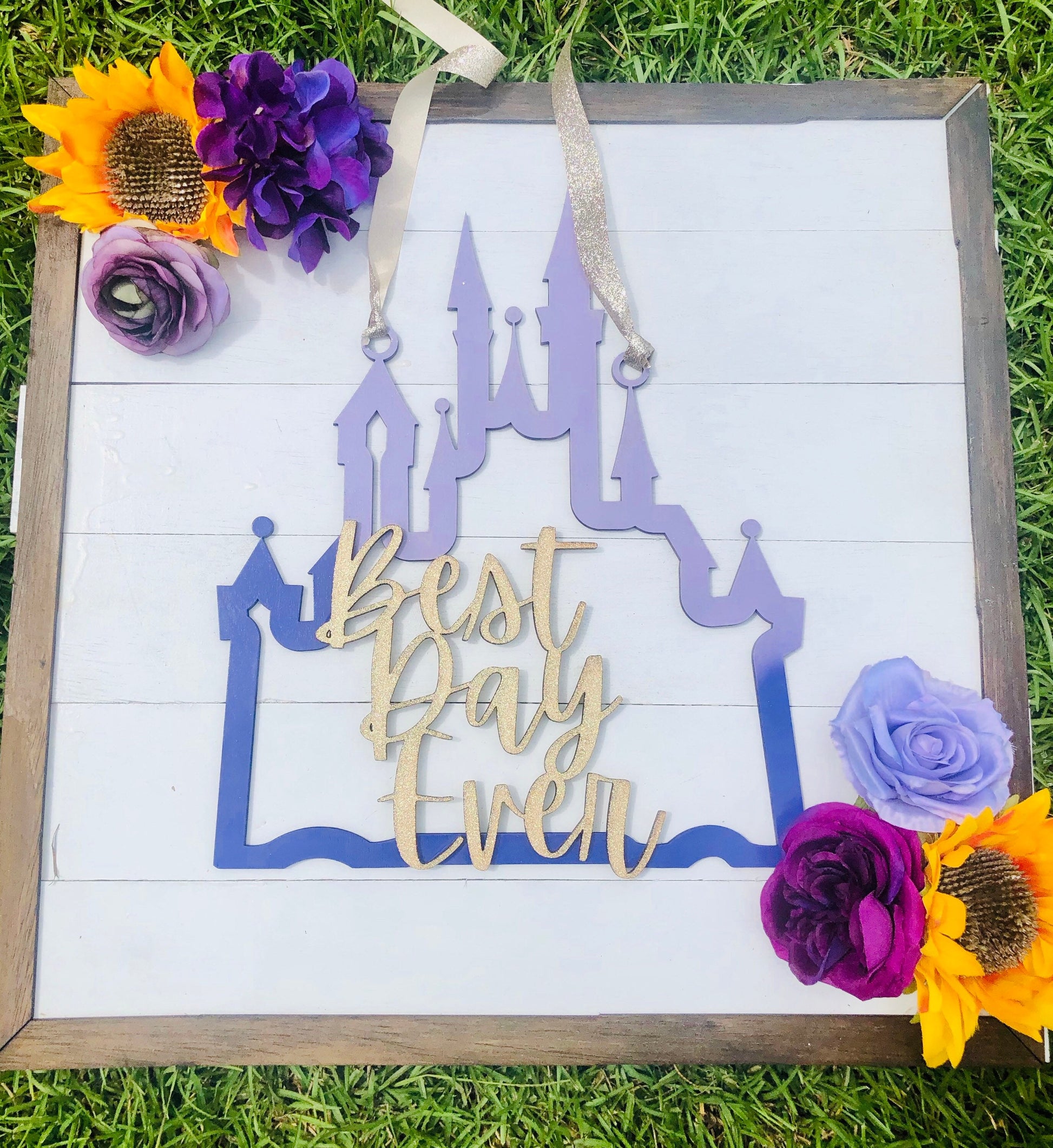 Decorative sign with castle design and 'Best Day Ever' text, surrounded by flowers on grass.