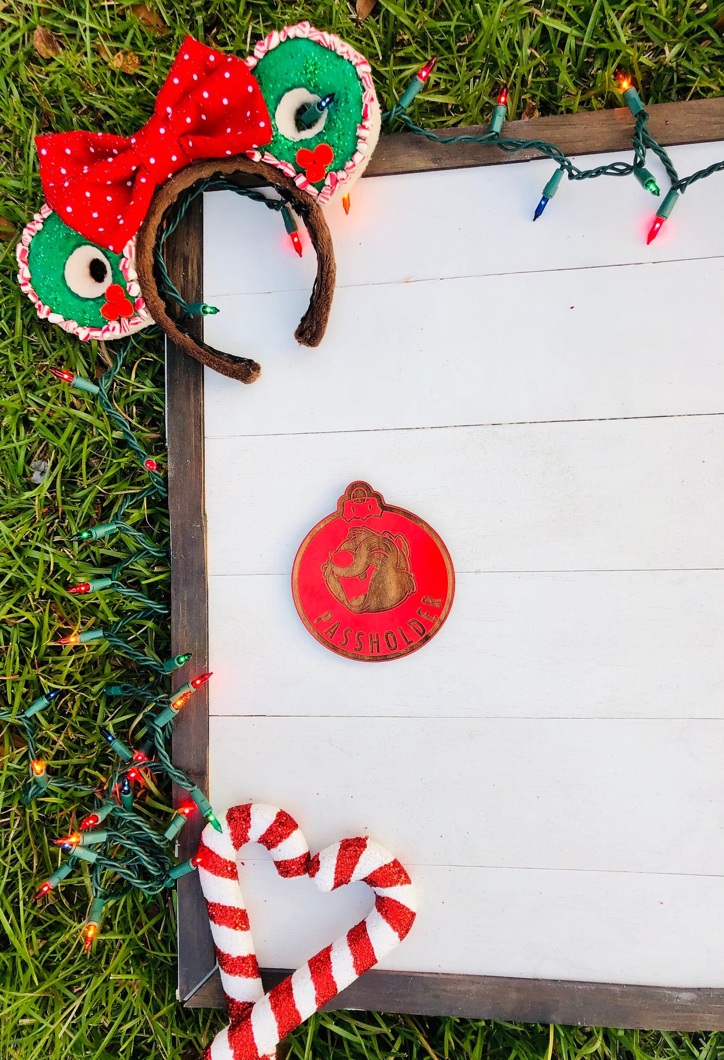 Decorative headbands with red bows and green accents, a candy cane, and a wooden ornament on a white surface with grass and lights in the background.