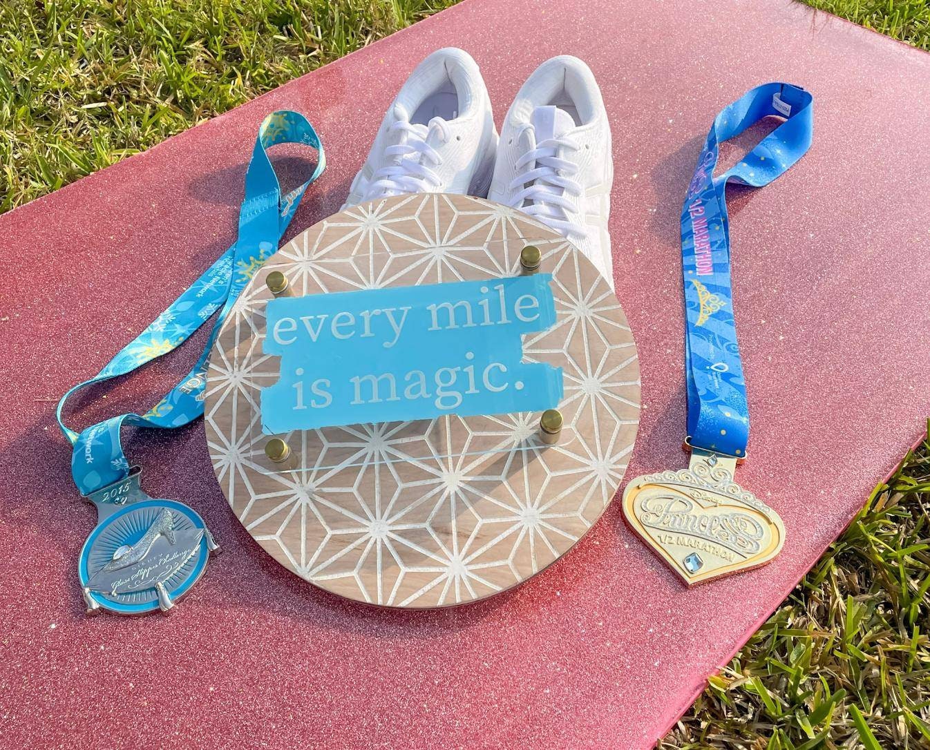 White sneakers on a pink mat with two medals and a sign reading 'every mile is magic' on a grass background.