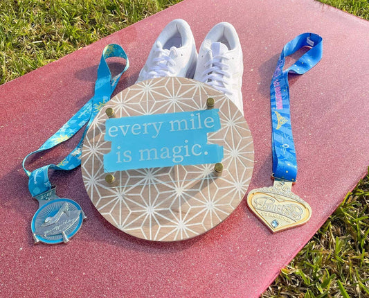 White sneakers on a pink mat with two medals and a sign reading 'every mile is magic' on a grass background.